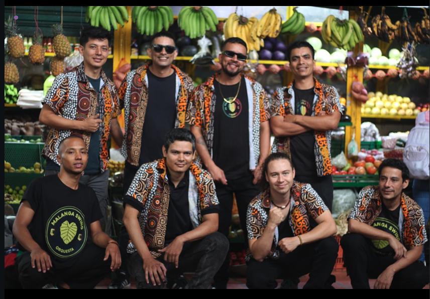 Foto de los nueve integrantes de Malanga Swing Orquesta en una plaza de mercado, con camisetas de la agrupación